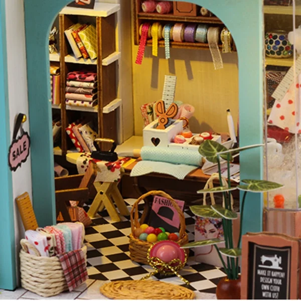 Close-up of the workbench inside the Tailor Shop miniature dollhouse kit, featuring sewing tools, fabric rolls, and design sketches.