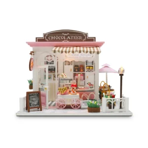 A miniature model of a chocolate dessert shop named "Cocoa Fantasy", featuring a striped awning, a blackboard sign, a chocolate vending cart, and an intricately detailed interior with dessert displays and gift boxes. A pink parasol covers an outdoor seating area beside the shop.