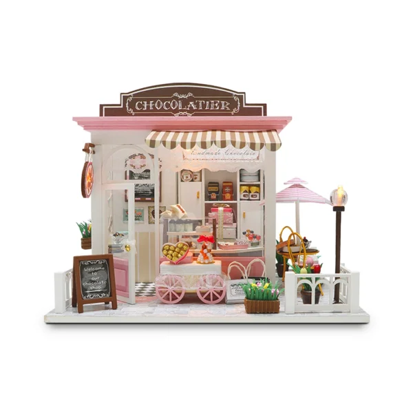 A miniature model of a chocolate dessert shop named "Cocoa Fantasy", featuring a striped awning, a blackboard sign, a chocolate vending cart, and an intricately detailed interior with dessert displays and gift boxes. A pink parasol covers an outdoor seating area beside the shop.