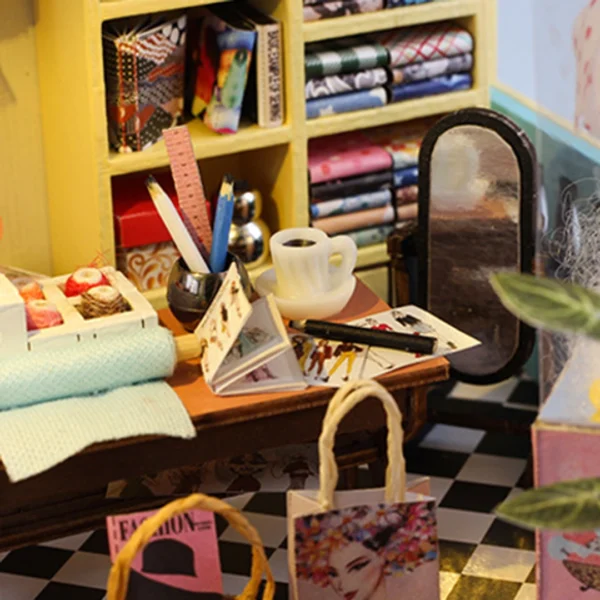 Close-up of a miniature tailor shop’s workbench, displaying sewing tools, design sketches, a coffee cup, and colorful fabric rolls in a cozy setting.