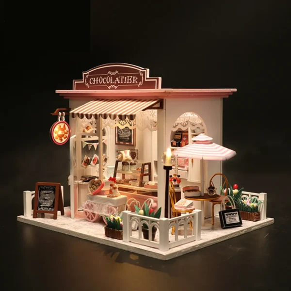 Side view of the Cocoa Fantasy Chocolate Shop, showing the storefront with detailed signage, an outdoor seating area, and soft lighting that enhances the miniature dessert shop's warm and inviting atmosphere.